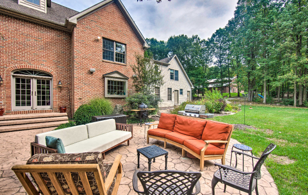 Outdoor patio surrounded by trees, with lots of patio furniture and grill, and the brick-exterior home in the background. 