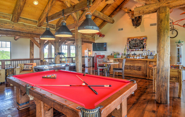 Image of a pool table with a red top inside a large rustic game room with wooden beams and pendant lights. A wet bar, wall decor, chairs, air hockey, and foosball table are in the background  